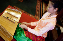 Asian woman dressed ornately kneeling on floor while playing  stringed instrument.