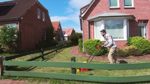Young man cutting the grass with the lawn mower in front of the house next to a fence