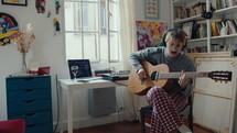 Young musician in headphones sitting on chair at home, playing the guitar and creating the music. Audio recording equipment on desk in background

