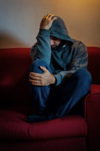 Young man is sitting alone on a red couch, holding his head in his hands, suffering from depression