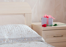 Gift box and a long-stemmed red rose on a nightstand by a bed.