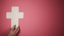 A hand placing a wooden cross on a pink background 
