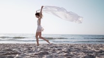  Slim beautiful woman running on sea beach with white silk fabric fluttering in wind.Сoncept of tenderness, lightness, art and talent in nature.

