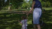 a mother pushing a stroller and holding her toddler daughter's hand walking in a park 