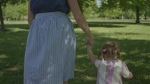 a mother and toddler girl walking in a park holding hands 