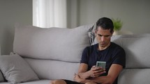 Middle aged male sitting on couch in living room and using his smartphone in leisure time. Mid shot of man chilling, browsing his phone and chatting.
