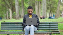Focused man freelancer sitting bench typing on laptop close up. Business black man resolving work issues using computer in green park. Serious man doing remote job.

