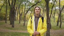 Cheerful young man in yellow coat with backpack standing and smiling in forest.