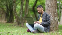 Young student on the grass in the park sits under a tree and cheerfully works with a laptop
