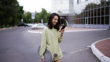 A brunette girl in a green sweater walks in the park. The girl has a skateboard in one hand and a phone in the other. The girl is texting on the phone. Evening walk.