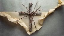 rustic wood cross with thorny stems and a background of torn and crumpled manila paper on a mottled gray surface