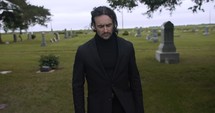 Sombre, sad young man in black suit walking in cinematic slow motion in cemetery through graveyard tombstones.