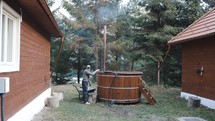 Man Putting Wood Into Furnace Of A Traditional Wooden Hot Tub Outdoor. wide