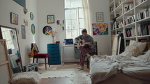 Young talented guitarist sitting by desk with audio equipment, playing the guitar, creating music at home recording studio in cozy room with unmade bed, bookshelves and DIY interior. Zoom Shot
