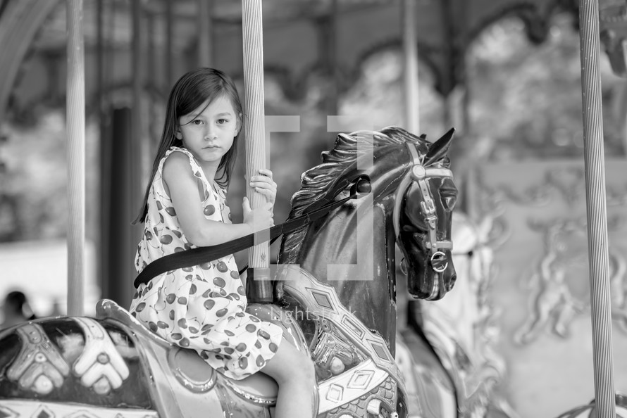 Black and white photo of a young girl in a cherry dress riding on a carousel 