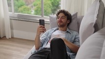 Satisfied caucasian guy is enjoying watching videos on couch. Carefree calm white man watch videos on phone, resting on sofa at home 
