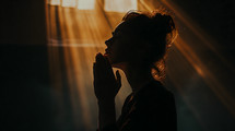 A woman praying with folded hands near a window with light coming through
