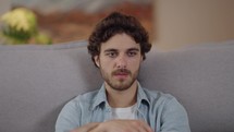 Portrait of young adult man sitting on the couch with serious worried sad expression worries about the past 
