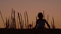 A girl on a beach crafts a bamboo barrier around herself, observing a colorful wind spinner in the sand, all amidst the soft glow of a summer sunset