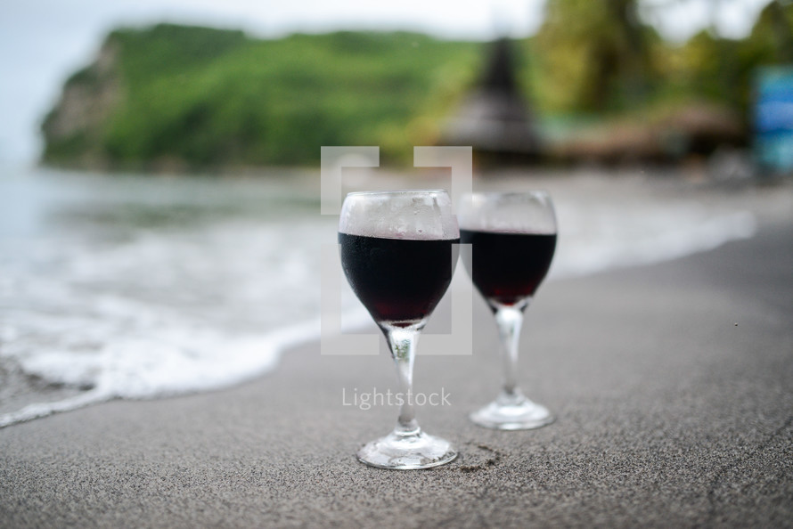 Two glasses of wine at the edge of the water on a beach.