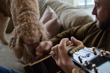 man sitting on a couch playing a guitar next to his dog