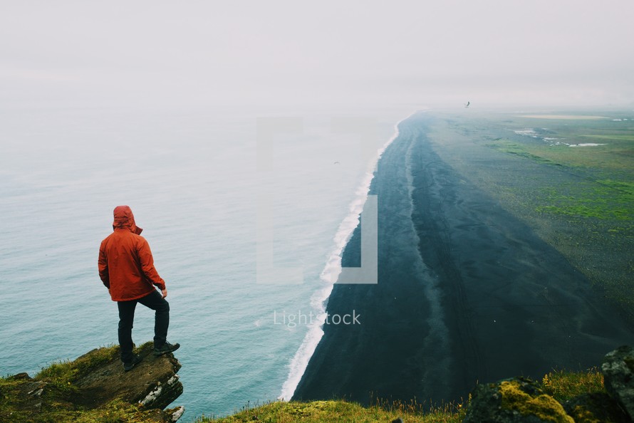A man standing on a cliff edge above the ocean at a black sand beach by the Dyrhólaey lighthouse in Iceland