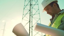 Worker male engineer using documents for checking data while standing against high voltage power towers. Power engineering specialist with working documentation working near electric poles.