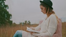 a woman sitting in a chair reading a Bible in a field 