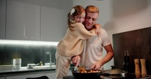 Happy little girl with braided hairstyle in a cream sweater together with her dad a middle-aged man with gray hair and wearing a white T-shirt prepare dinner in a modern kitchen before their dinner in the evening