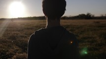 Young man, teenage boy standing in a field alone and watching the sunset or sunrise in cinematic slow motion.