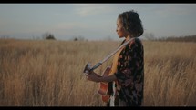 a woman standing in a field holding a guitar singing 