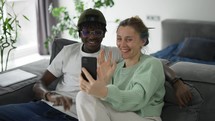 Happy biracial couple sitting on sofa, and having video call on smartphone.