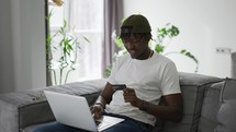 Young black man holds plastic bank card, sitting on a comfortable sofa, shopping using a laptop.