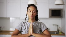 Young African woman doing meditation at home, connecting hands in namaste.