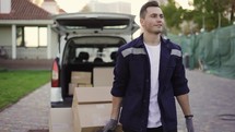 Young handsome smiling Caucasian delivery man with boxes on cart going from van with opened truck. Male post office worker Courier concept.