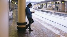 man reading a Bible leaning against a column near train tracks at a train station 