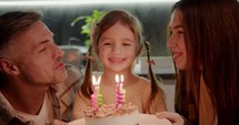 Close-up portrait of a little girl with a braided hairstyle in a cream sweater blowing out four candles on her birthday cake in a modern kitchen with her parents