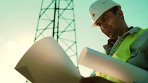 Worker male engineer using documents for checking data while standing against high voltage power towers. Power engineering specialist with working documentation working near electric poles.