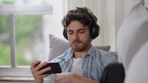 Caucasian man in wireless headphones holds smartphone choose podcast for listening use audio digital streaming media services provider resting on sofa spend leisure at home. Wireless, modern tech
