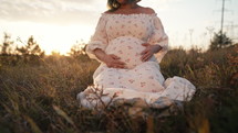 Happy Mother Carrying Child, Expecting For Baby. Amazing Motherhood. Pregnant Woman in beautiful dress sitting on grass at nature rural meadow. High quality 4k footage 