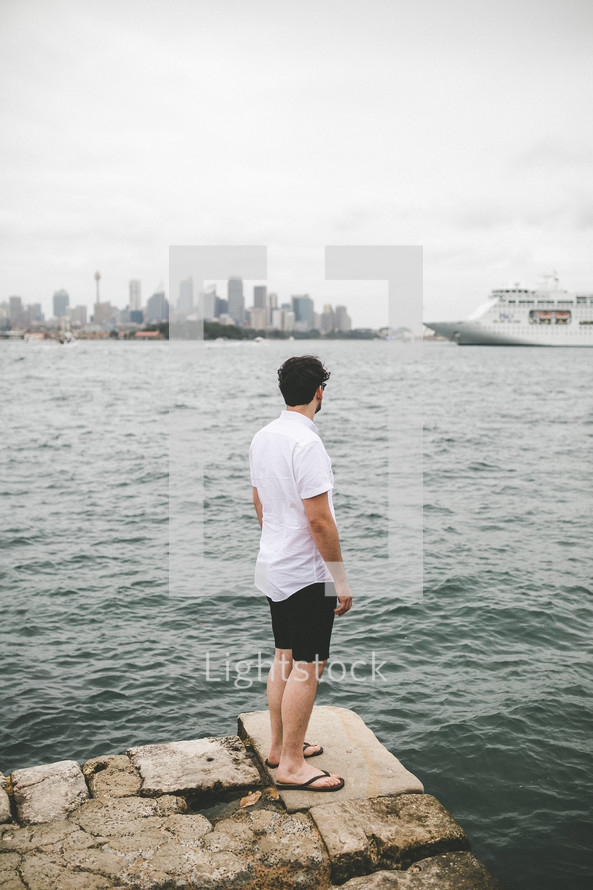 a man standing on a rocky shore watching a passing cruise ship 