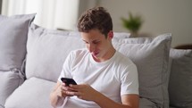 Happy caucasian teen boy holding cell phone using smartphone device at home. Smiling young brazilian man blogger subscribing new social media, buying in internet, ordering products online in apps.
