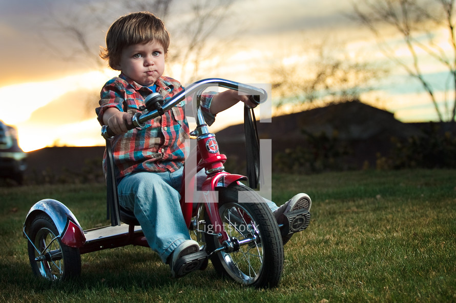 Toddler boy riding red tricycle on grass with mountains and trees in background at sunset.