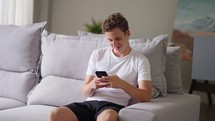 Caucasian teenager is playing with mobile phone in the living room on the sofa. Happy young handsome guy sitting on couch, Smiling enjoy communicating with friends or social network at new
