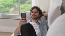 Portrait of young man resting on sofa with a phone. Guy is laying on a couch uses mobile phone. Student studying online through phone at home.
