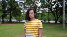 Unhappy male person face outdoors portrait. Looking at camera close up. Upset shy guy. Kind people portraits. Young adult hippie at a park. Hipster hairstyle.
