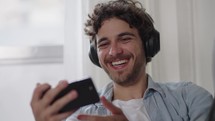 Cheerful young caucasia man watch funny video and laughing wearing headphone. Funny male layinug on the couch at home having fun listening podcast 
