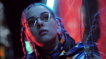 Young pretty girl with unusual hairstyle near glowing red neon lights of the city at night. Dyed blue hair in braids. hipster teenager in glasses 