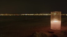 A candle in a glass jar prominently aglow on a nighttime beach, with the lights of a distant coastal city softly blurred in the background.