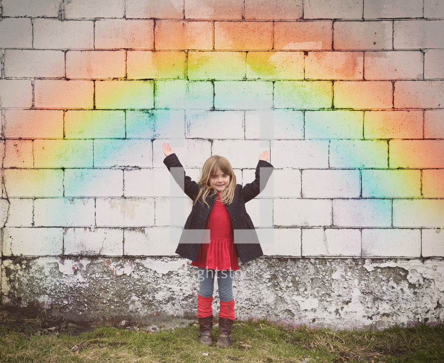a little girl with raised hands standing under a rainbow painted on a wall 